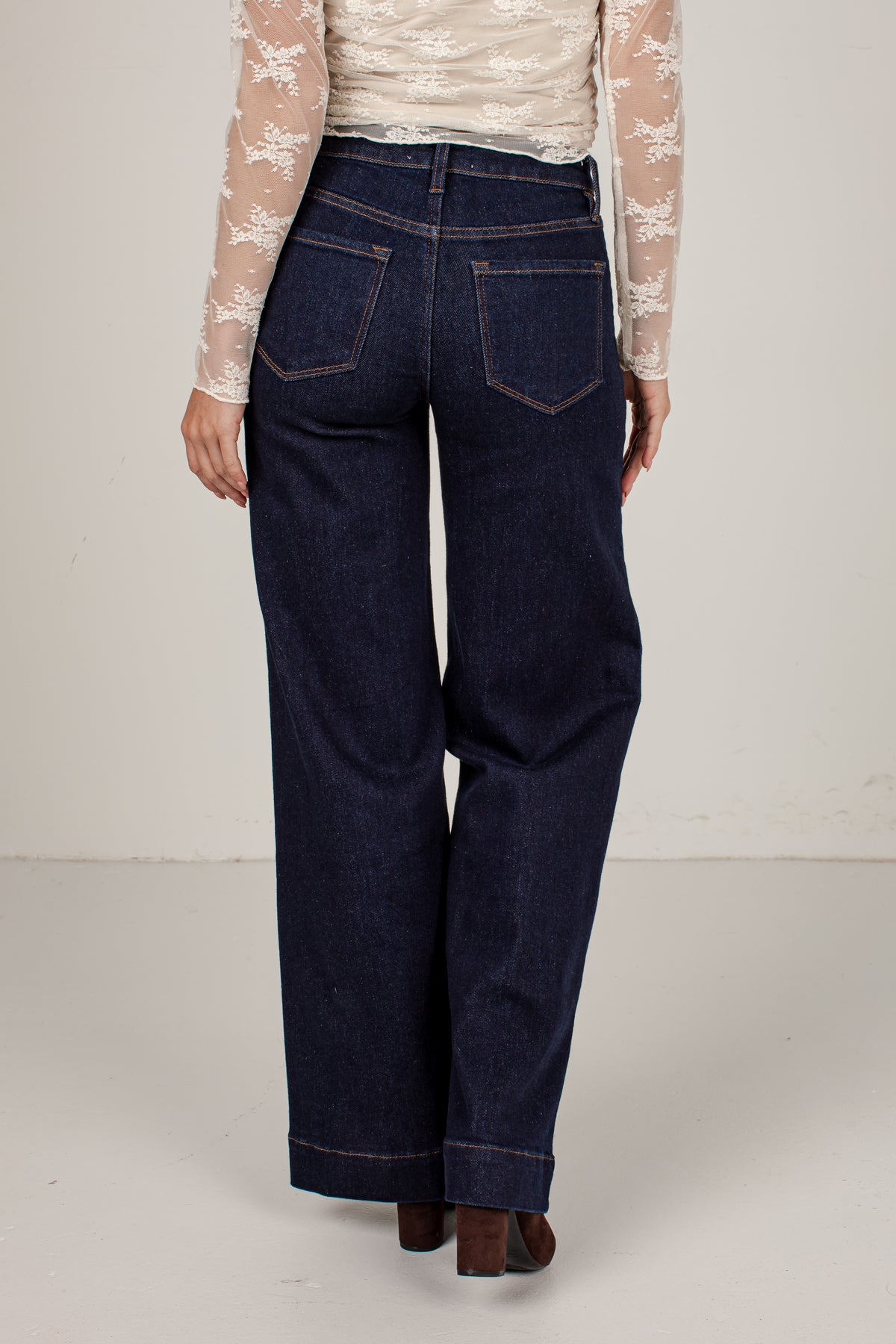 A person shown from the back wearing dark indigo wide-leg jeans, a cream lace long-sleeve top, and brown heels against a plain off-white studio background.