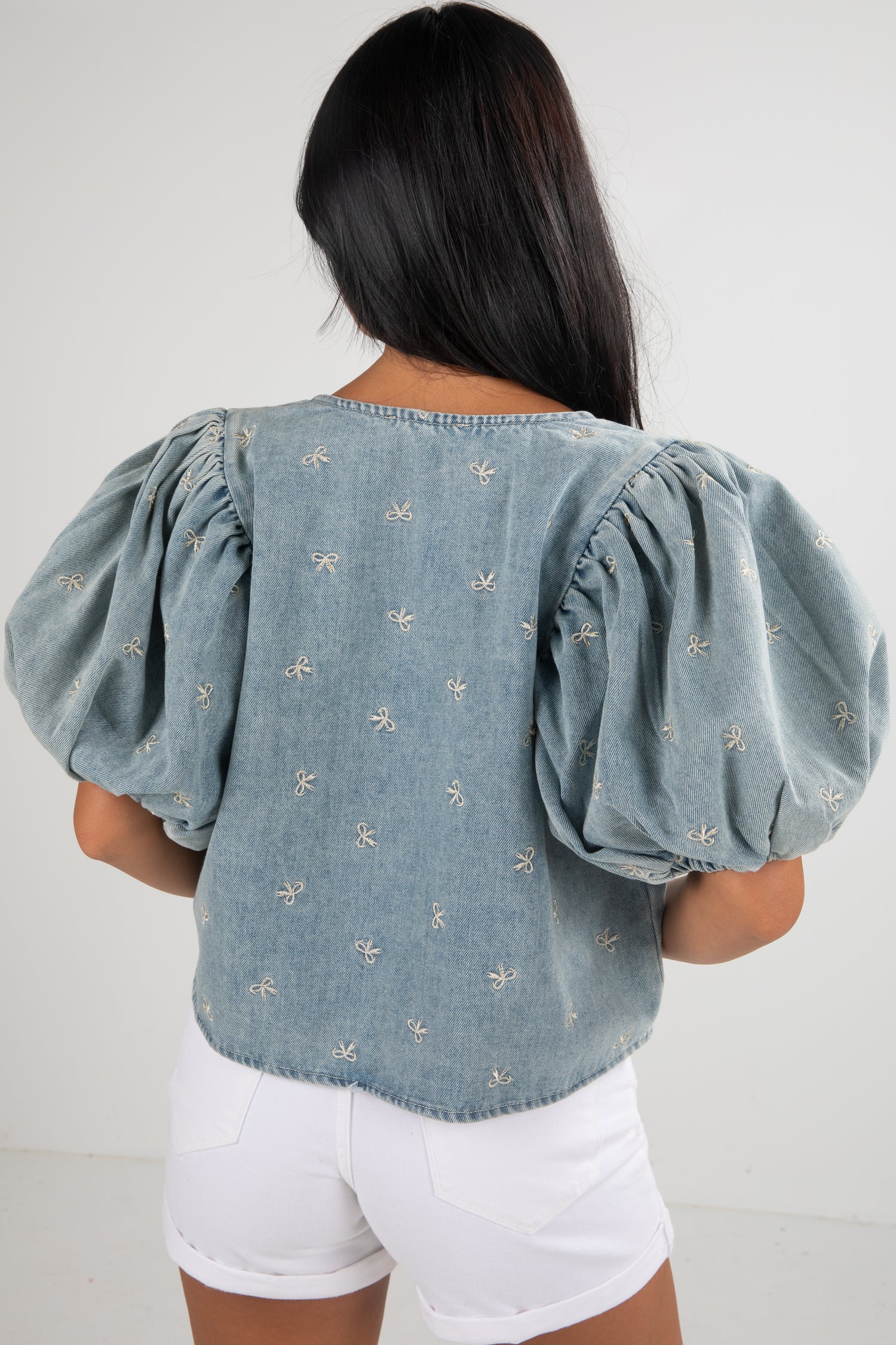 Woman wearing a light blue denim top with puff sleeves and white shorts on a plain background