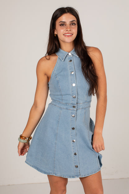 A woman modeling a sleeveless light blue denim halter mini dress with a button-down front, photographed against a plain light gray studio background.