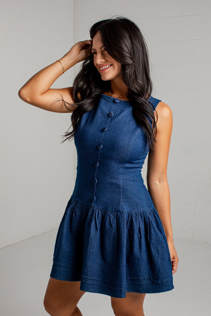 A woman modeling a sleeveless blue denim dress with a buttoned neckline and a flared skirt, standing against a light gray wall.