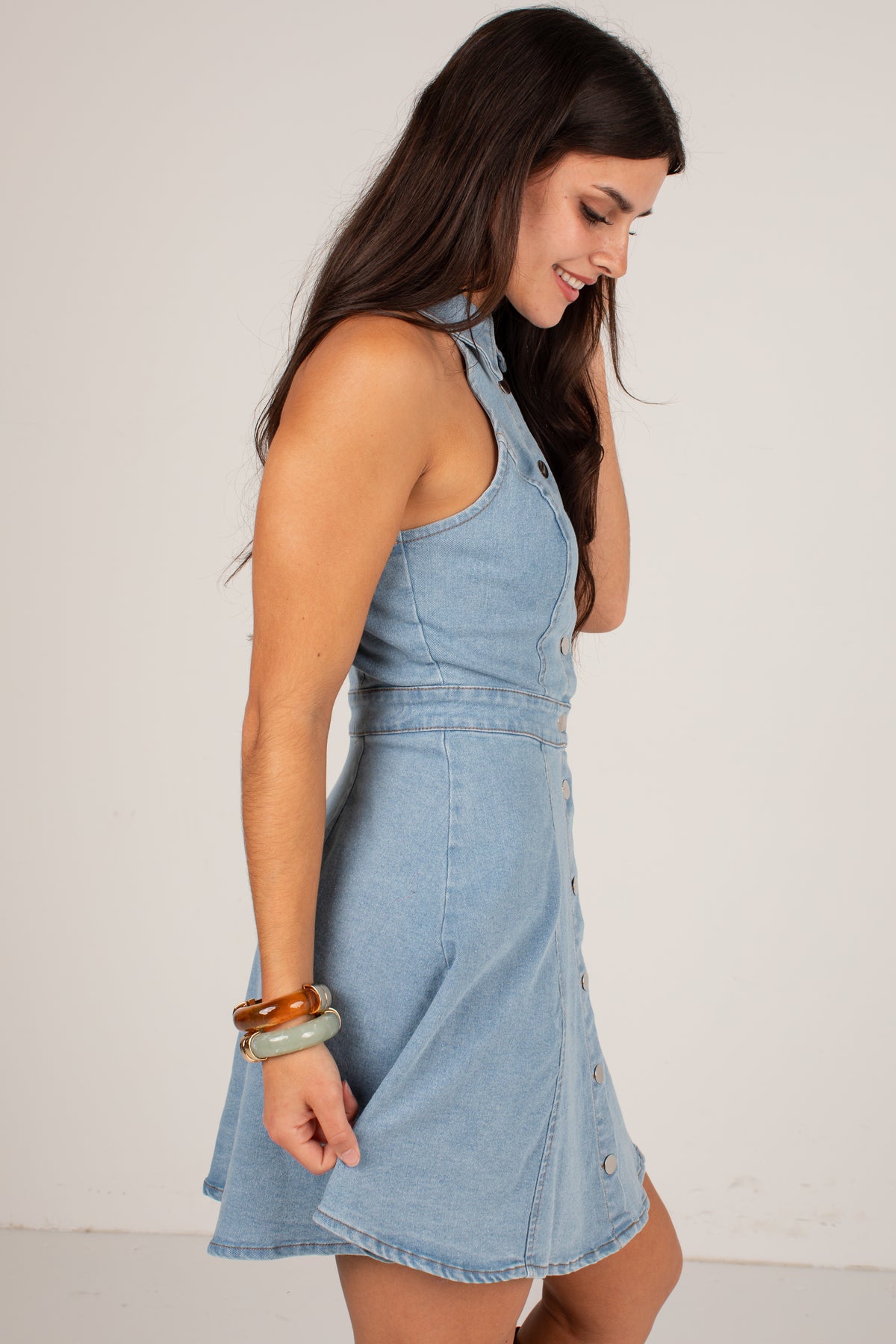 A woman wearing a sleeveless light blue denim mini dress, shown from a side profile against a plain off-white studio background.