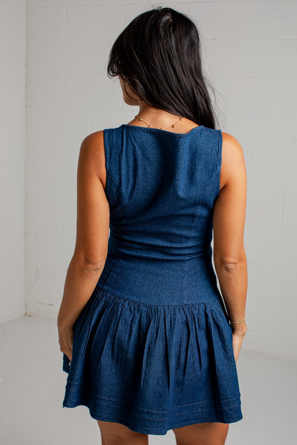 A woman viewed from behind wearing a sleeveless dark blue denim mini dress with a gathered waist and flared skirt, standing against a white wall.