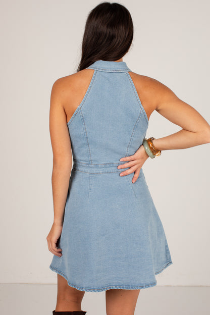 A woman shown from the back wearing a light blue denim halter dress, standing against a plain off-white studio background.