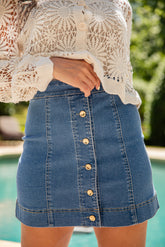 Person wearing a white lace top and blue denim skirt with buttons, standing by a pool.