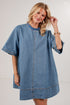 A smiling blonde woman wearing an oversized blue denim tunic dress with short wide sleeves, a round neckline, and contrast stitching, photographed against a plain light gray studio background.