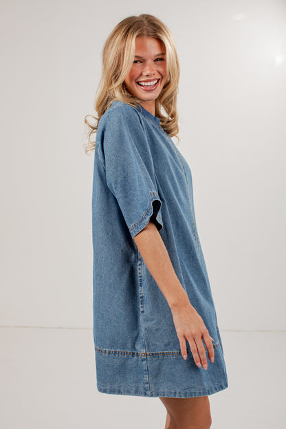 A smiling blonde woman wearing an oversized blue denim tunic dress, photographed against a plain light gray studio background.