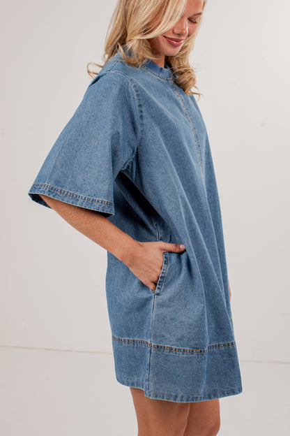 A woman wearing an oversized light blue denim dress with wide short sleeves, a high collar, and side pockets, shown against a plain off-white studio background.