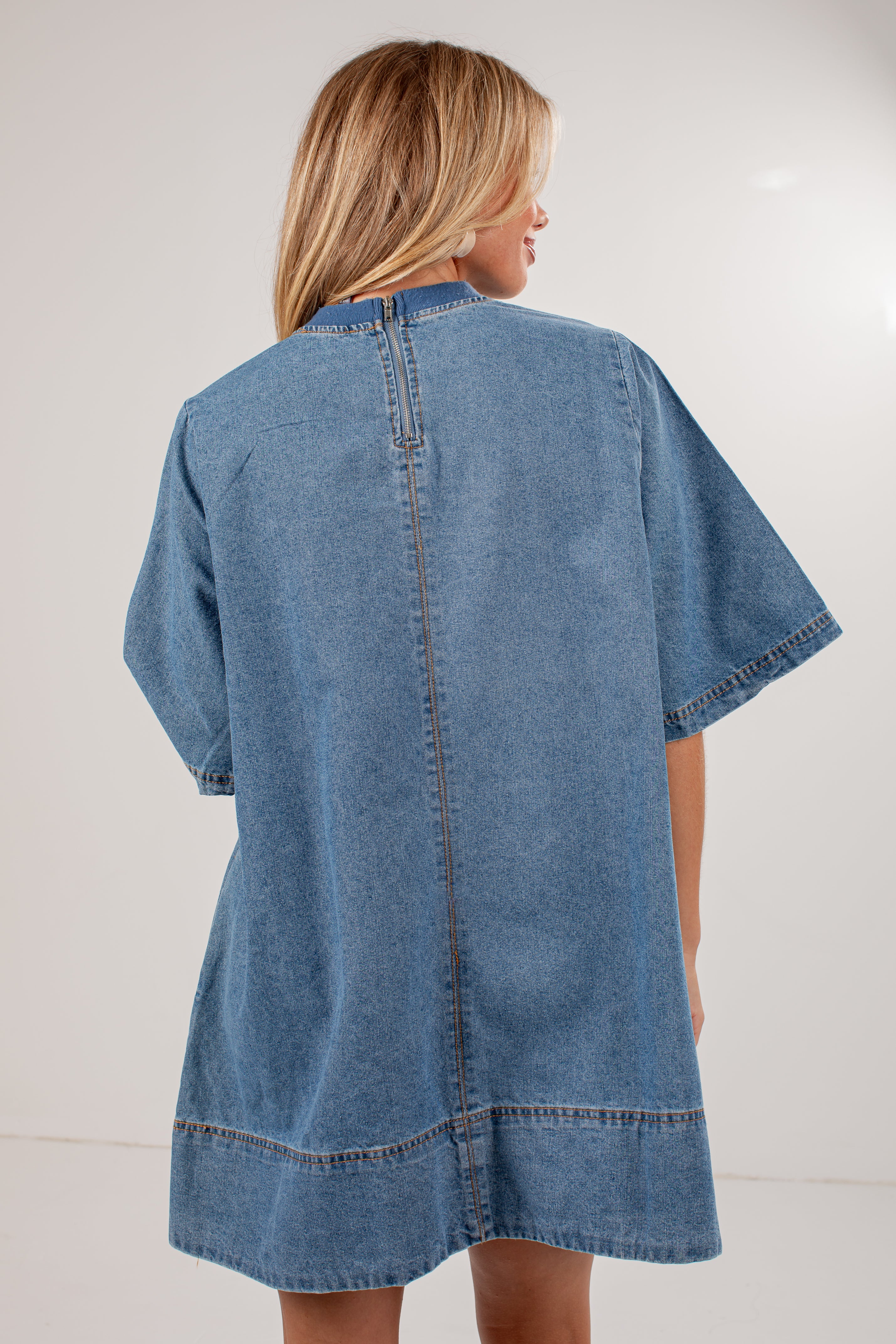 A woman shown from the back wearing an oversized light blue denim tunic or cover-up dress with short sleeves and contrast stitching, photographed against a plain light background.