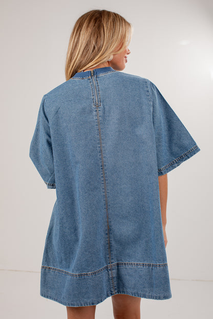 A woman shown from the back wearing an oversized light blue denim tunic or cover-up dress with short sleeves and contrast stitching, photographed against a plain light background.