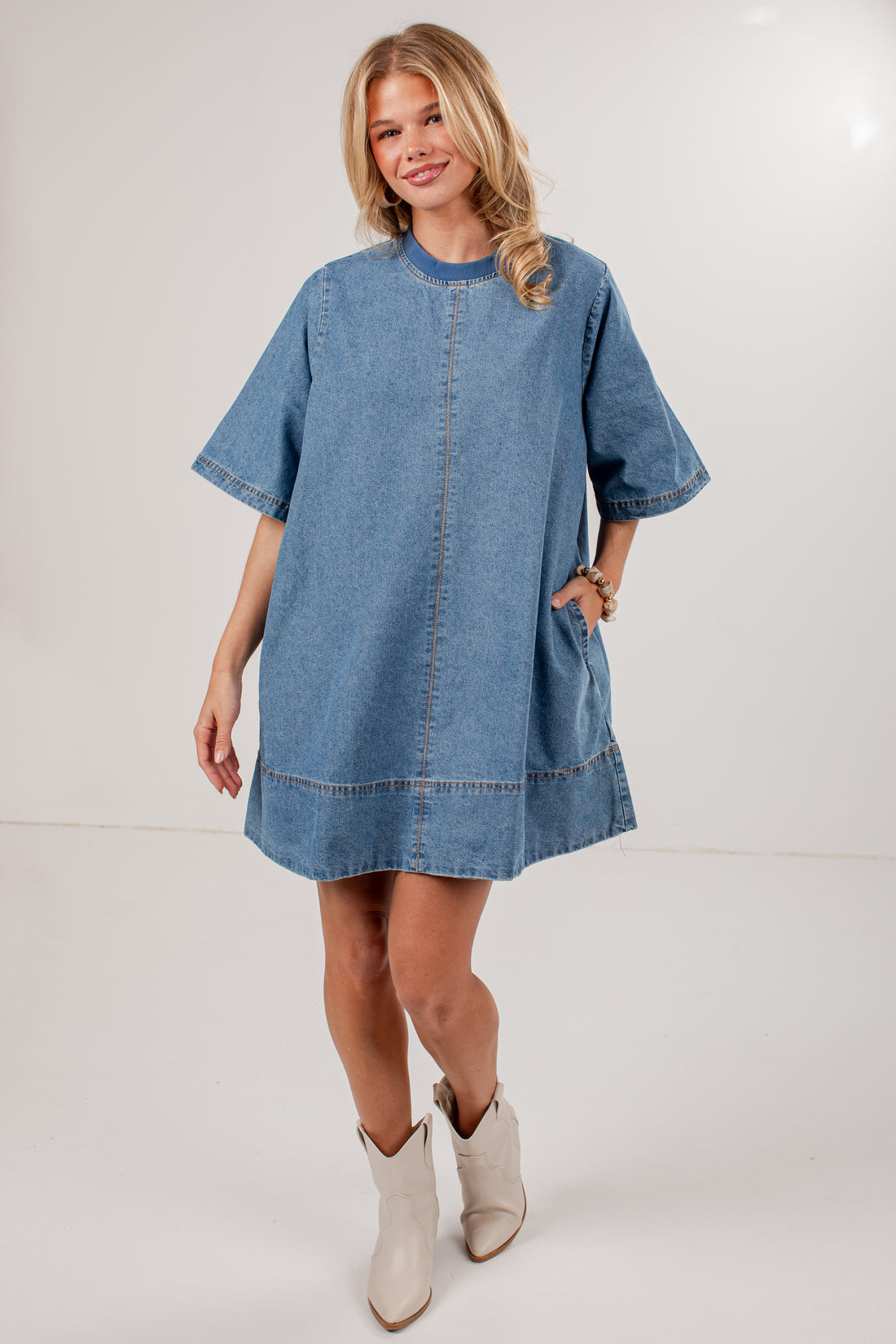 A woman modeling a loose-fitting blue denim dress with short sleeves, a round neckline, and front patch pockets, paired with light beige ankle boots, against a plain light gray studio background.
