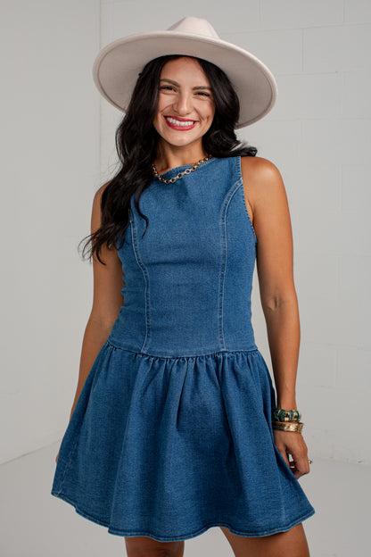 A woman wearing a sleeveless blue denim dress with a flared skirt and a cream wide-brim hat, standing against a light gray wall.