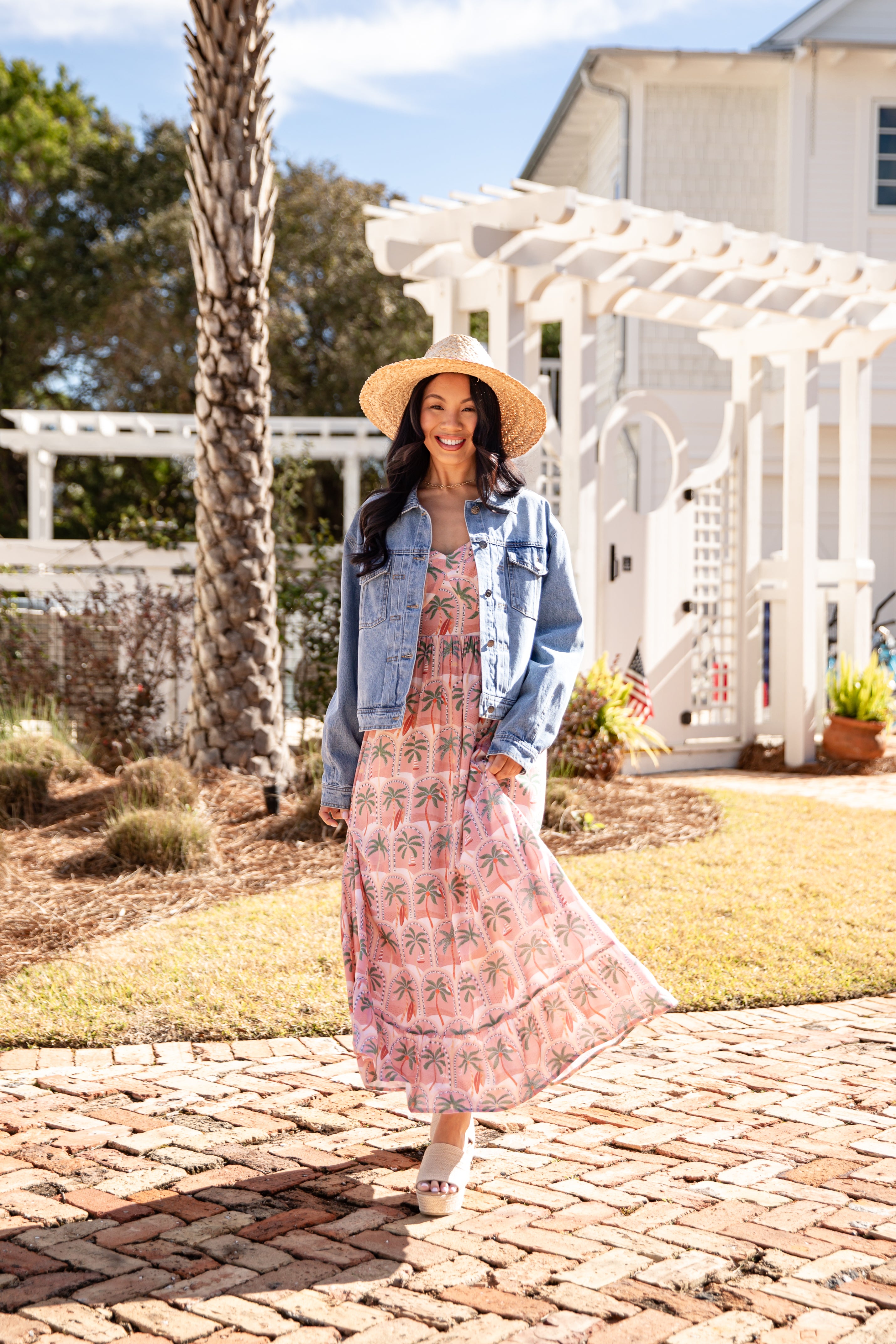 Woman in a floral dress and denim jacket standing on a brick path with a white building and trees in the background.
