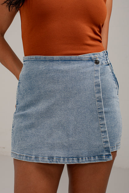 A light blue denim wrap mini skirt worn by a model, paired with a ribbed orange-brown tank top, shown from the chest down against a neutral light gray background.