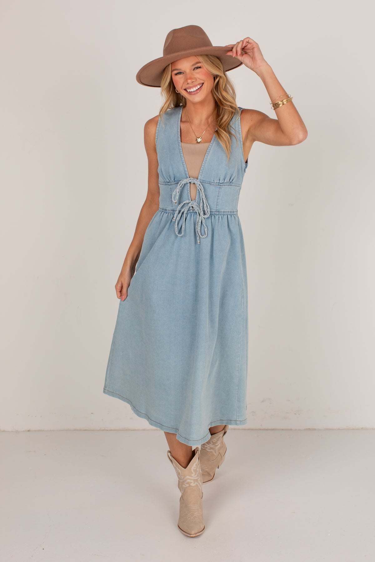 A woman modeling a sleeveless light blue denim midi dress with a tie-front detail, styled with a brown wide-brim hat and beige ankle boots against a plain off-white studio background.