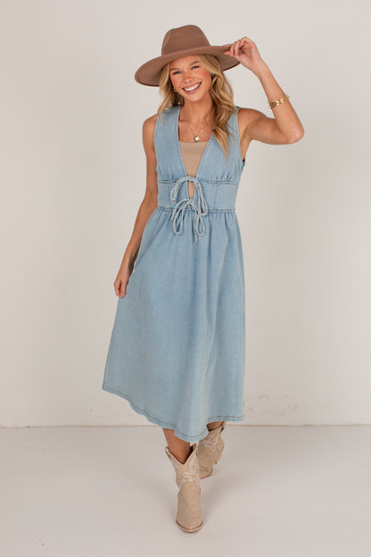 A woman modeling a sleeveless light blue denim midi dress with a tie-front detail, styled with a brown wide-brim hat and beige ankle boots against a plain off-white studio background.