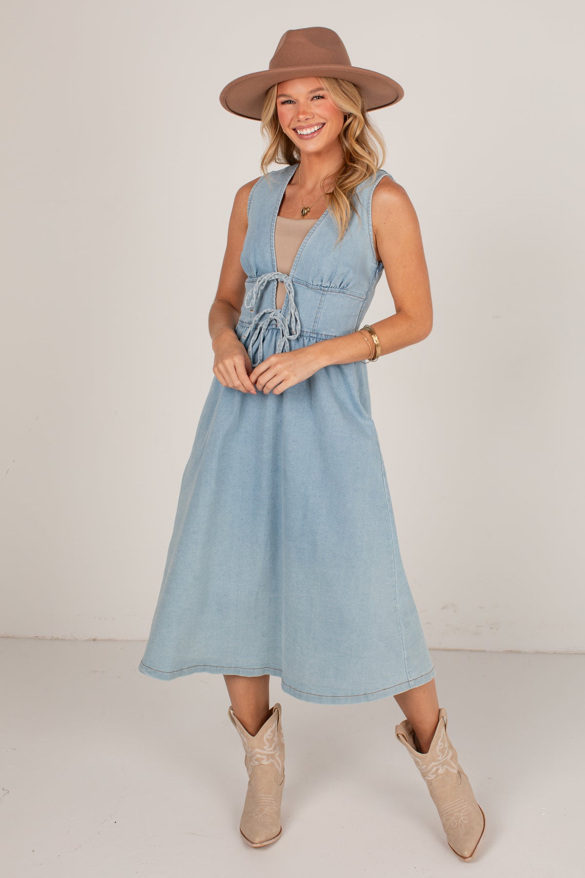 A woman modeling a light blue denim midi dress with a tie-front detail, styled with a brown wide-brim hat and beige cowboy boots against a plain studio backdrop.