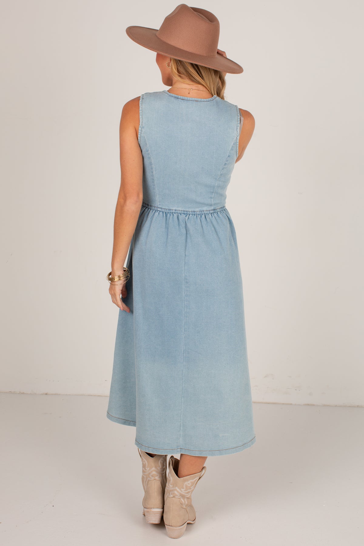 A woman shown from the back wearing a sleeveless light blue midi dress, a brown wide-brim hat, and beige cowboy boots, standing against a plain off-white studio background.