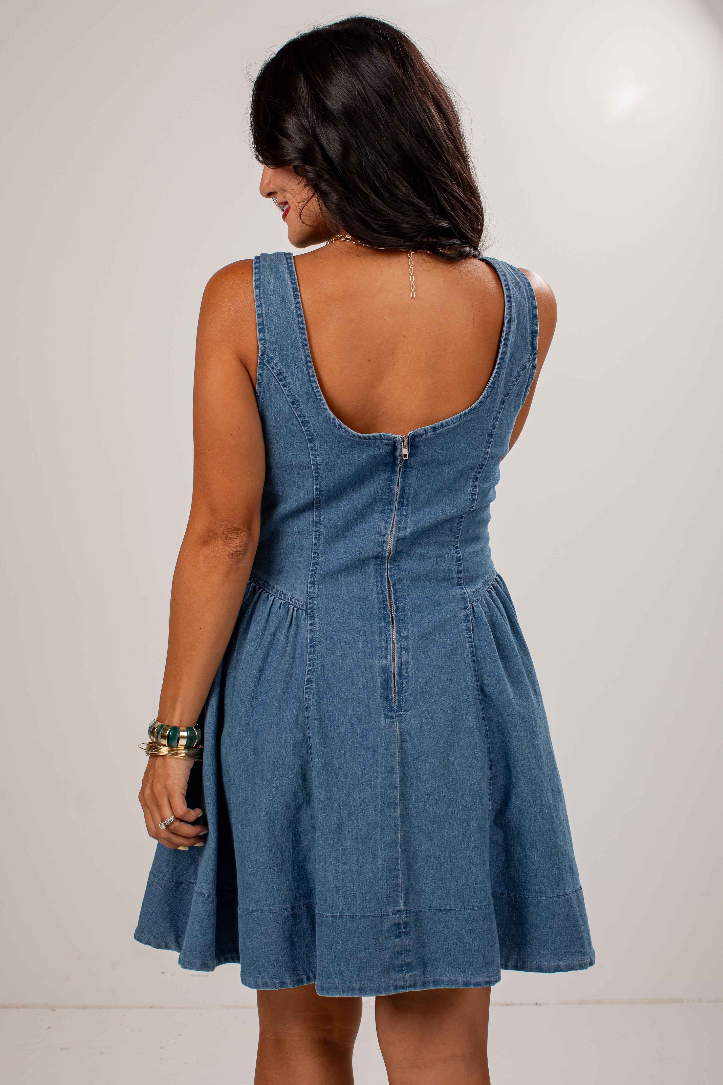 A woman shown from the back wearing a sleeveless blue denim dress with a wide scoop neckline, gathered waist, and flared skirt, photographed against a plain light background.