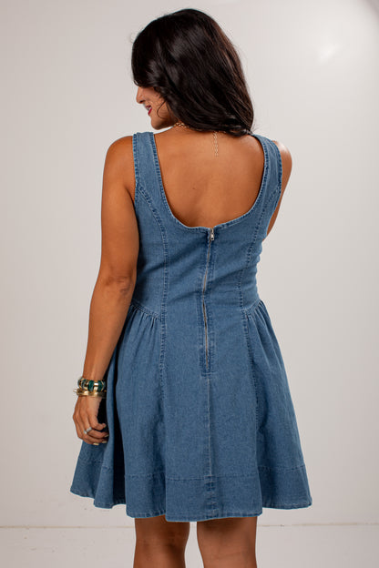 A woman shown from the back wearing a sleeveless blue denim dress with a wide scoop neckline, gathered waist, and flared skirt, photographed against a plain light background.