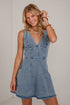 A woman modeling a sleeveless light blue denim mini dress with a V-neckline and button details on the bodice, standing against a white brick wall.