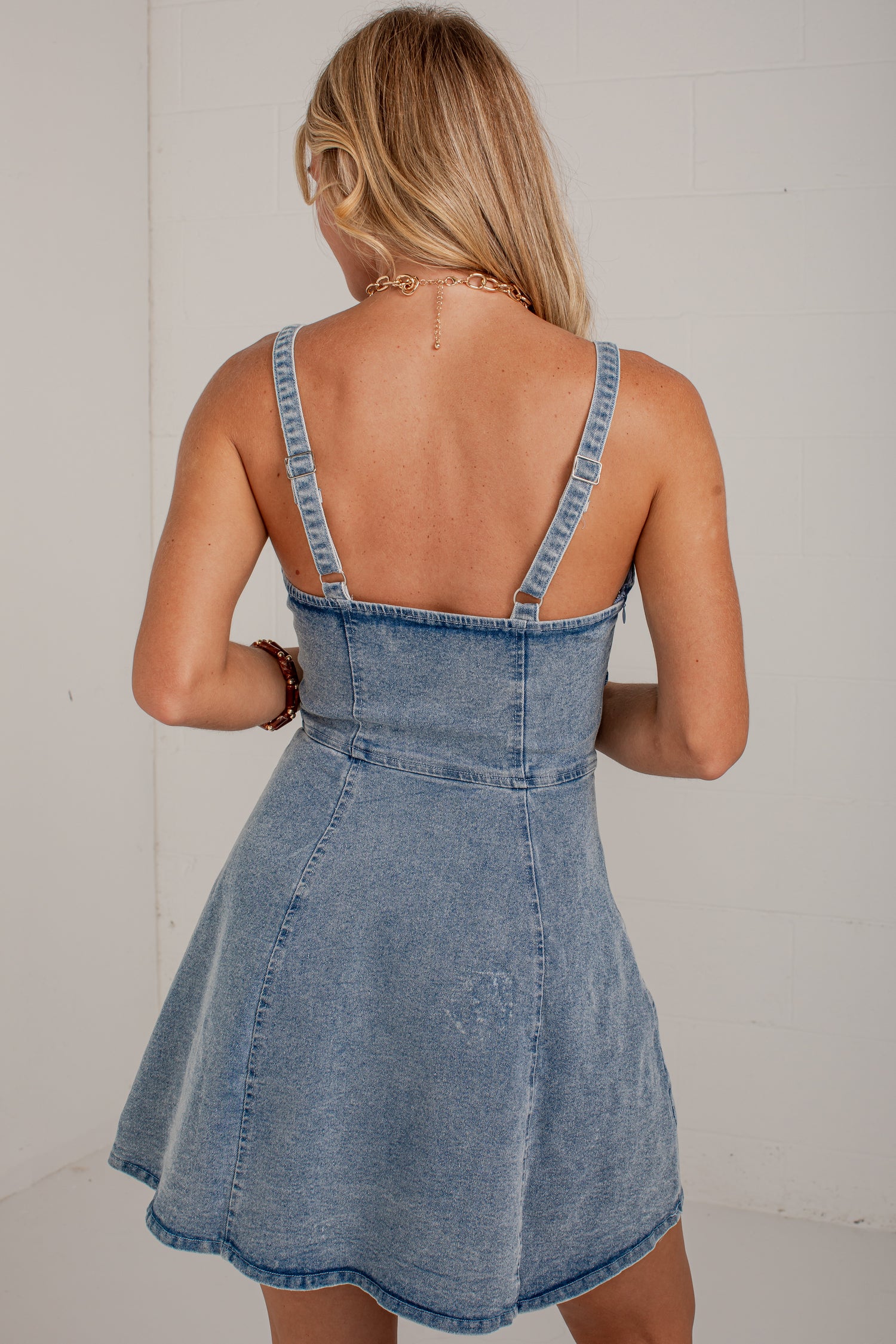 A woman viewed from behind wearing a light blue denim pinafore-style dress with thin shoulder straps and a flared skirt, standing against a plain white wall.