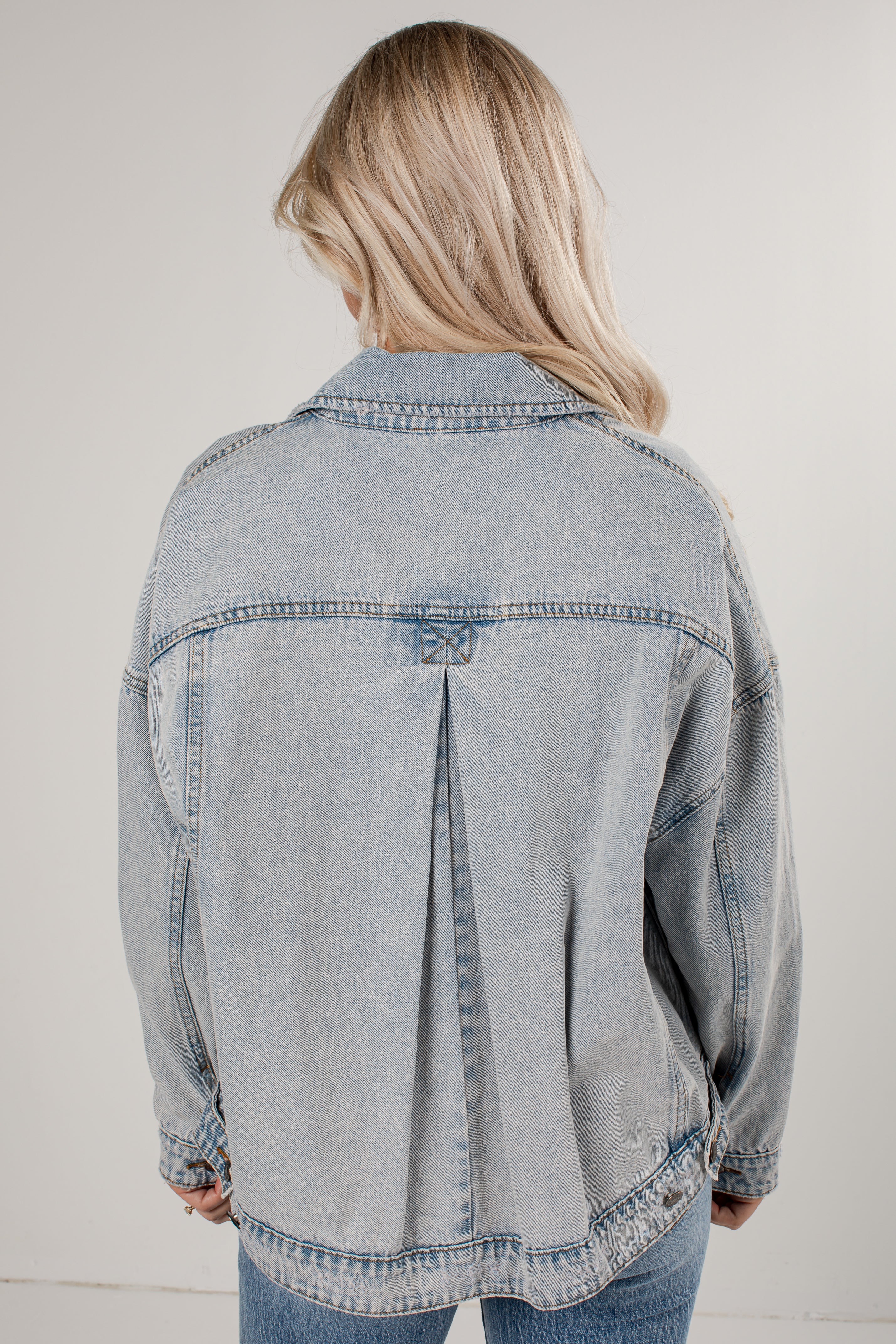 A woman viewed from behind wearing a light-wash blue denim jacket with a unique back seam detail, paired with blue jeans against a plain light gray background.