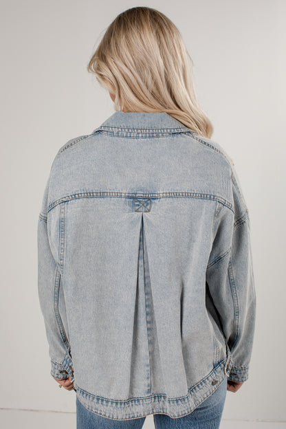 A woman viewed from behind wearing a light-wash blue denim jacket with a unique back seam detail, paired with blue jeans against a plain light gray background.