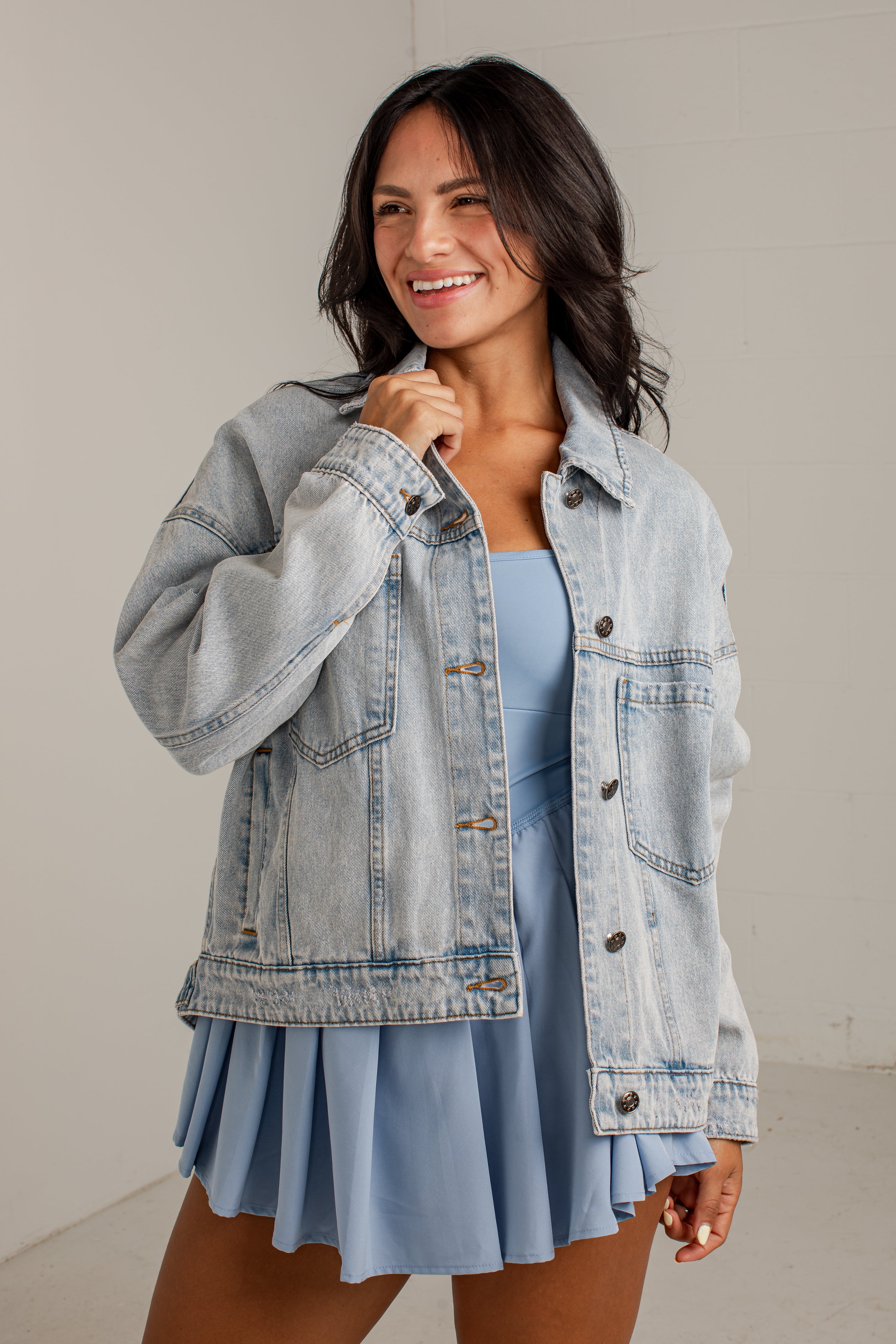 A woman wearing a light-wash blue denim jacket over a light blue pleated dress, standing against a neutral off-white wall.