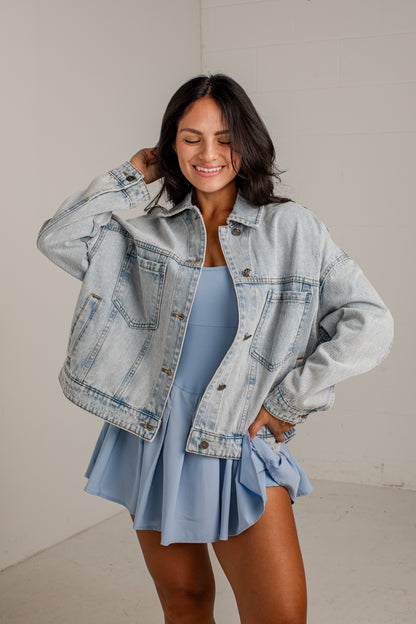A woman modeling a light-wash, oversized denim jacket over a light blue mini dress, standing against a neutral off-white studio background.