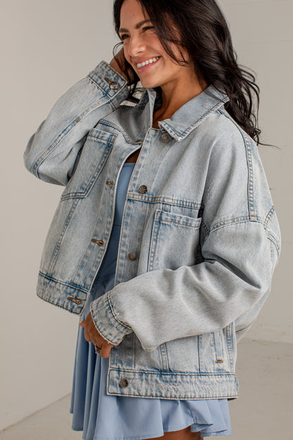 A woman wearing a light-wash blue denim jacket over a light blue dress, smiling and posing against a neutral beige studio background.