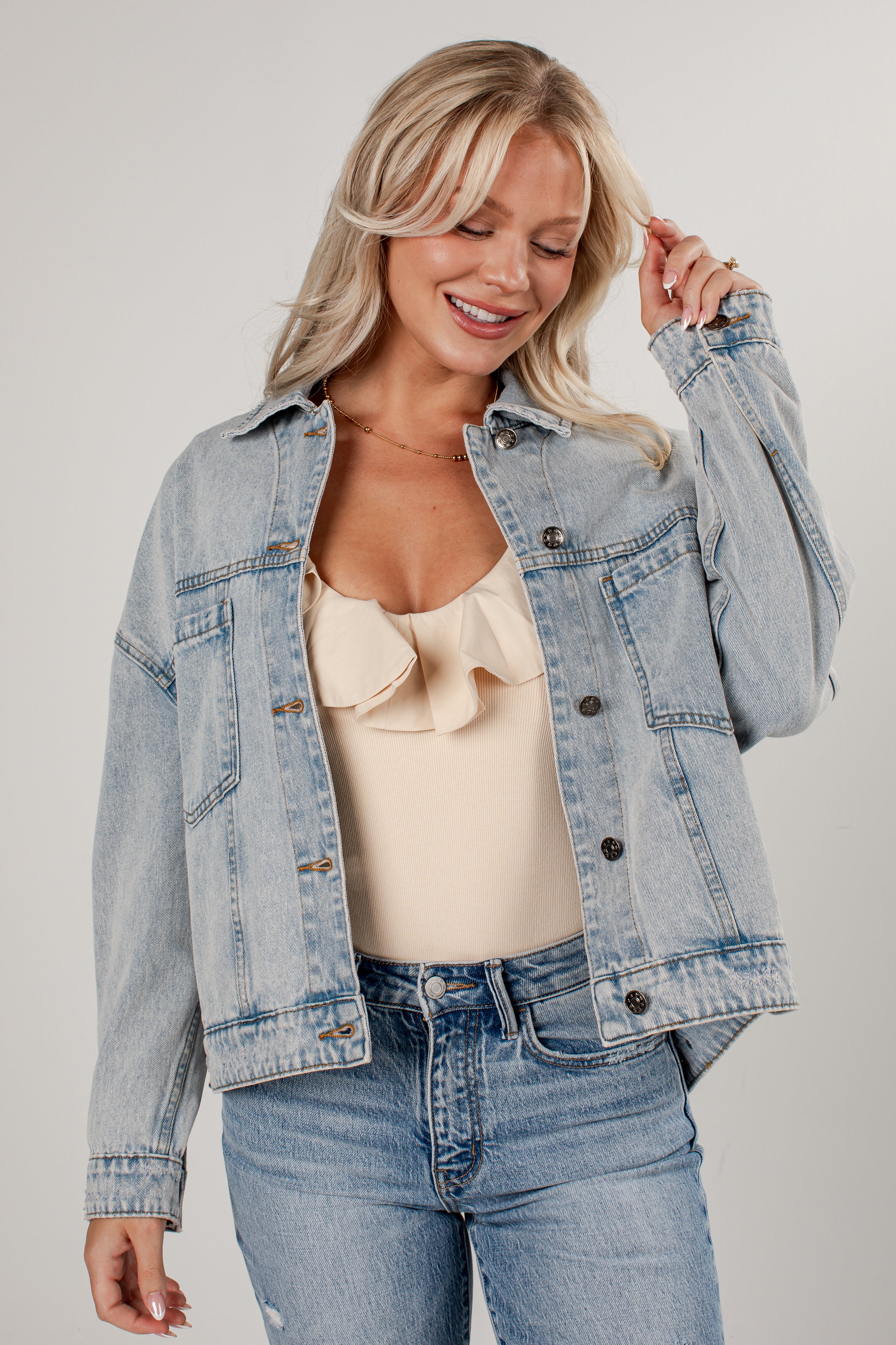 A woman wearing a light-wash blue denim jacket over a beige ruffled camisole top and blue jeans, posing against a neutral light gray studio background.
