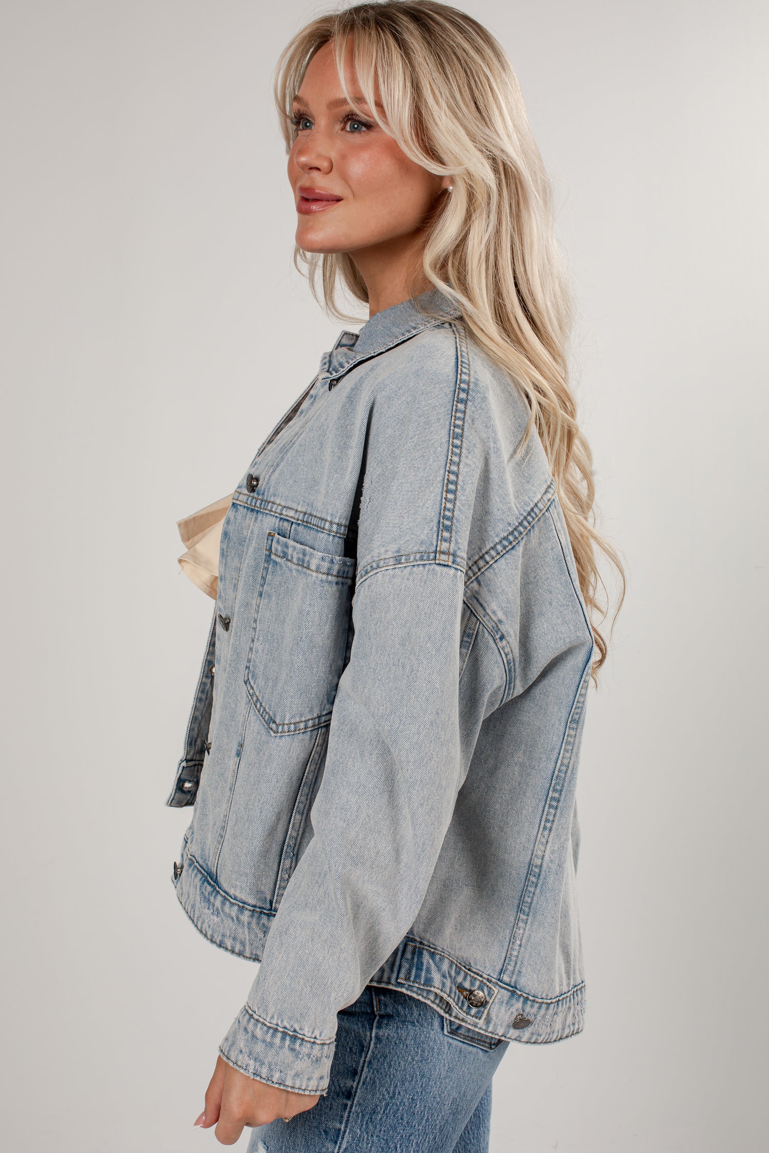 A woman wearing an oversized light-wash blue denim jacket, styled with a beige top and blue jeans, photographed against a neutral light gray studio background.