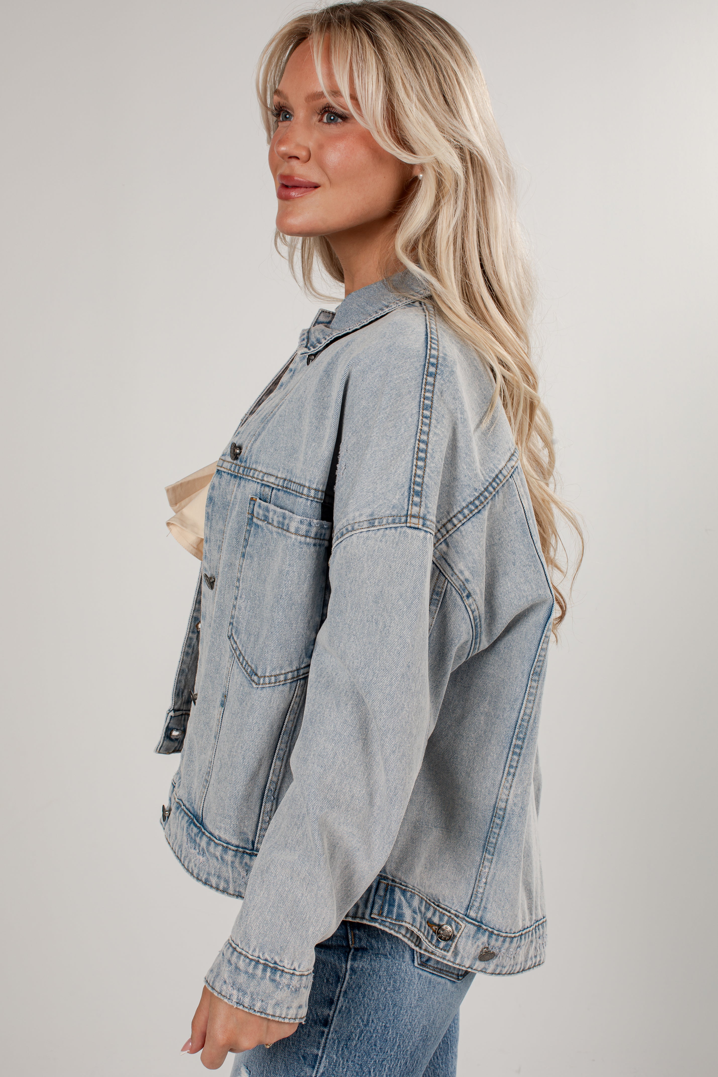 A woman wearing an oversized light-wash blue denim jacket, styled with a beige top and blue jeans, photographed against a neutral light gray studio background.