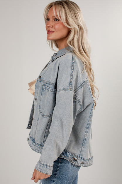 A woman wearing an oversized light-wash blue denim jacket, styled with a beige top and blue jeans, photographed against a neutral light gray studio background.