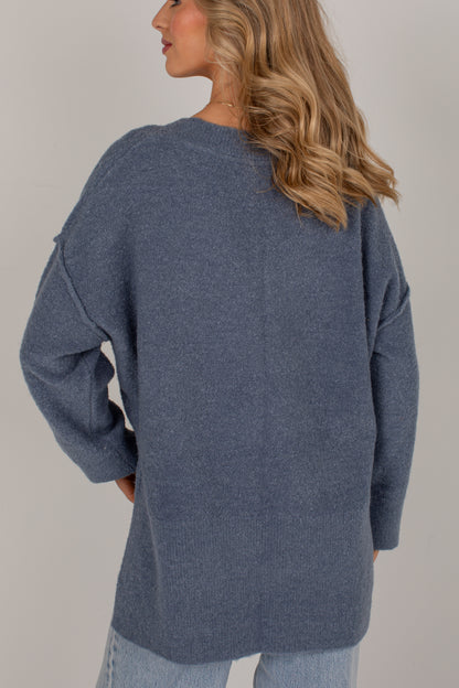 A woman wearing an oversized heathered slate blue knit sweater, shown from the back against a plain light gray studio background.