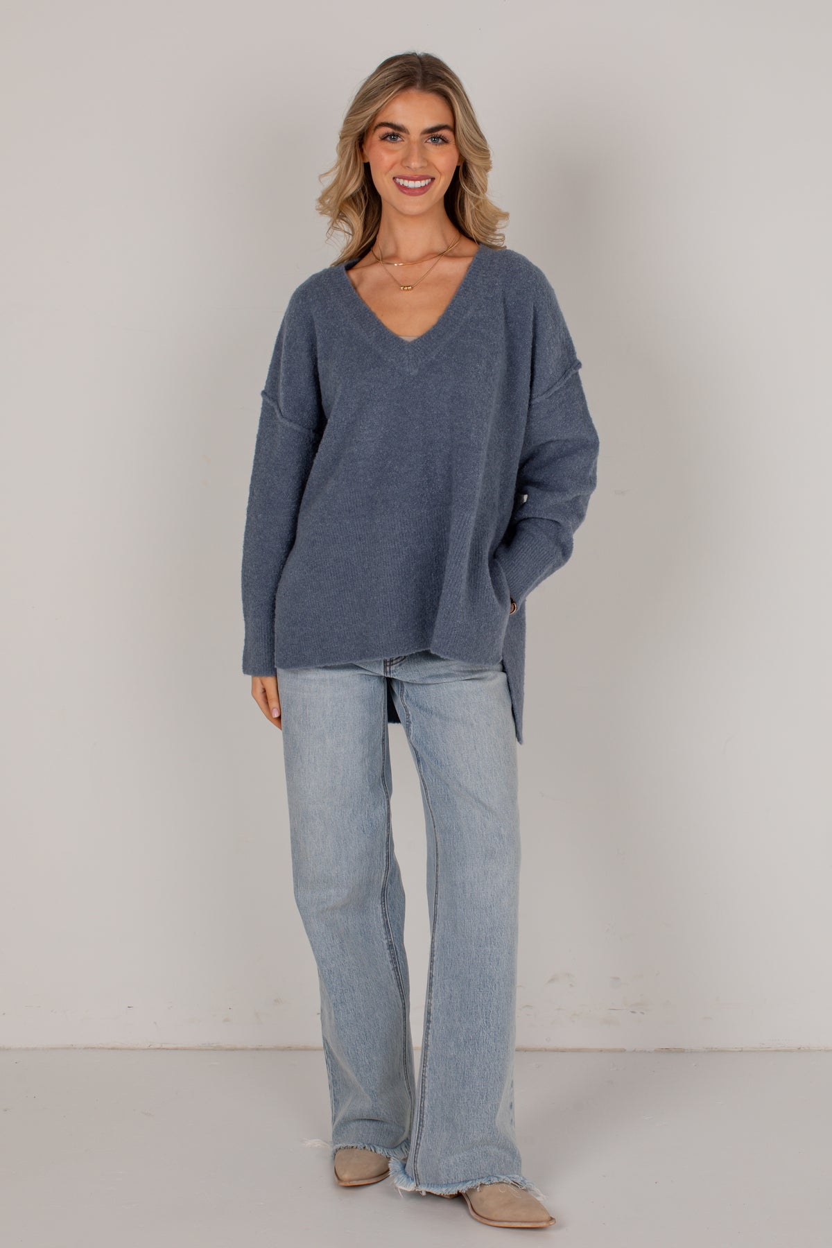 A woman modeling an oversized slate blue V-neck knit sweater with light-wash wide-leg jeans and beige flats, standing against a plain off-white studio background.