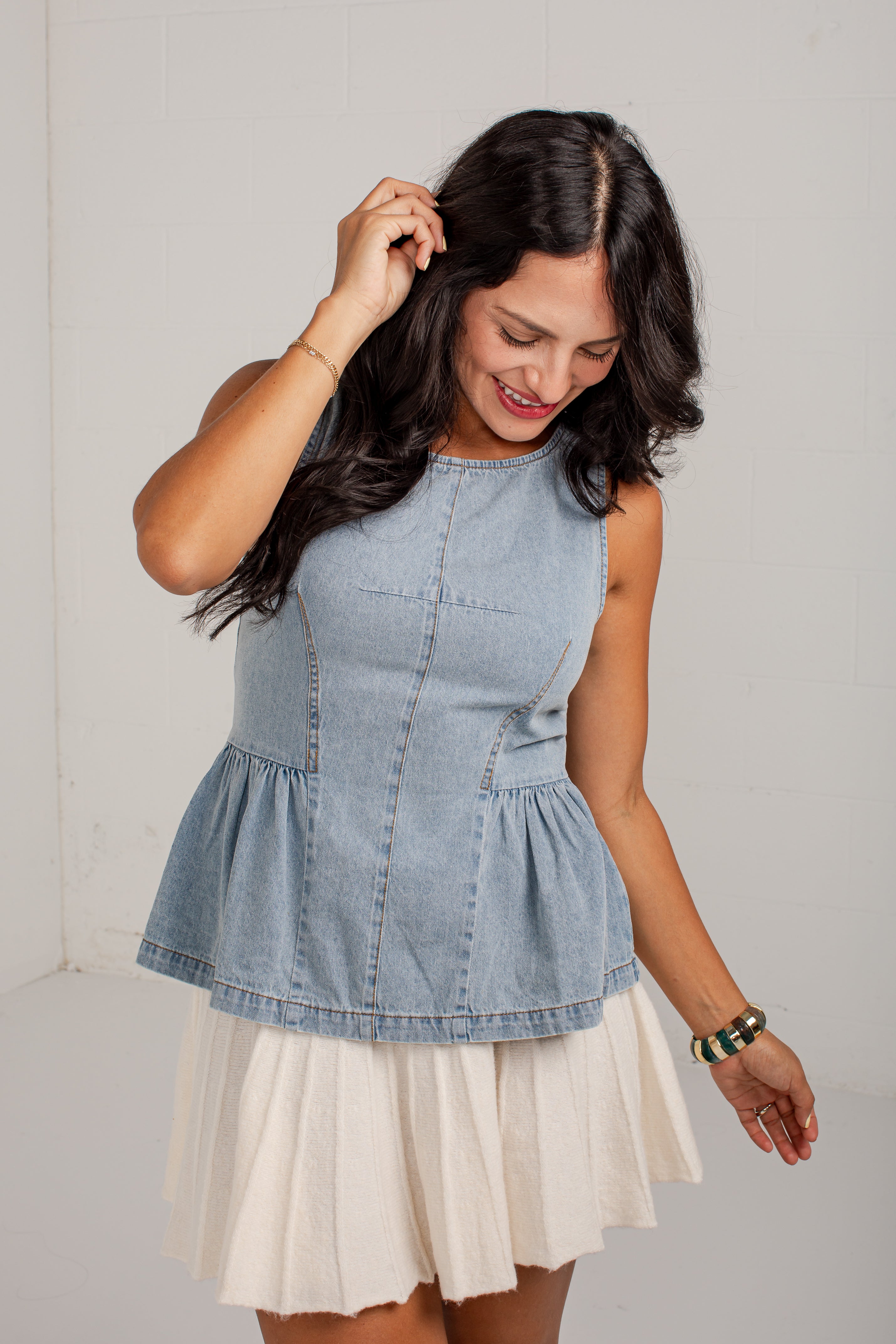A woman wearing a light blue denim sleeveless top with a peplum hem and a white pleated skirt, standing against a light gray wall.