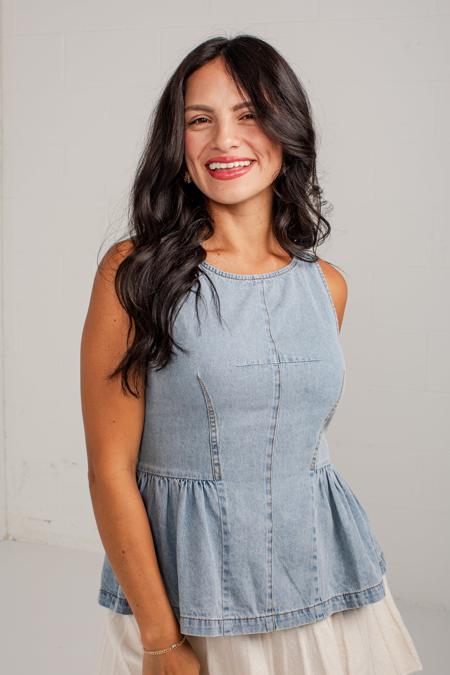 A smiling woman with long dark wavy hair wearing a light blue denim sleeveless top with a peplum hem and a white skirt, standing against a light gray wall.