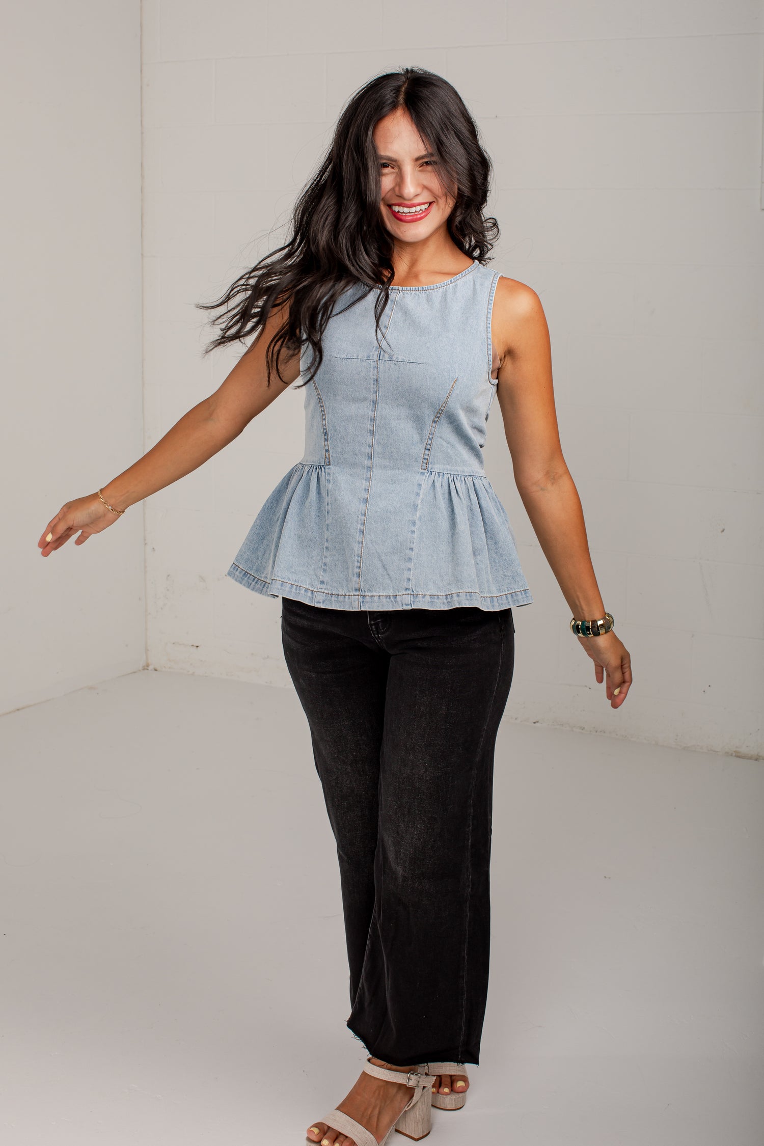 A woman modeling a light blue sleeveless denim peplum top with black wide-leg pants and beige sandals, standing in a white studio corner.