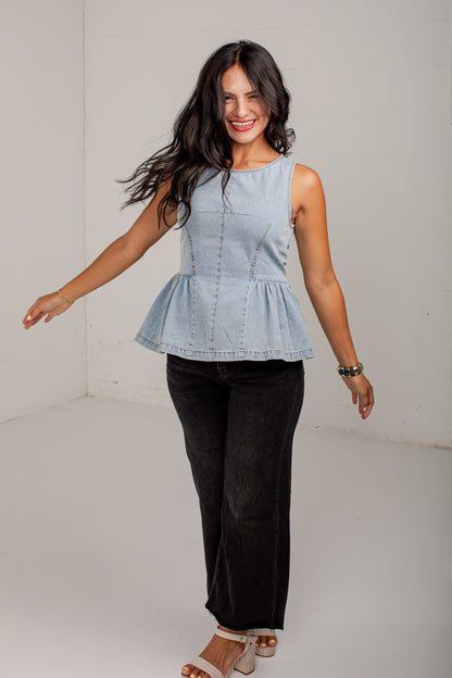 A woman modeling a light blue sleeveless denim peplum top with black wide-leg pants and beige sandals, standing in a white studio corner.
