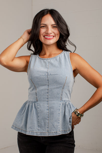 A woman modeling a light-wash denim sleeveless peplum top against a neutral light gray background.