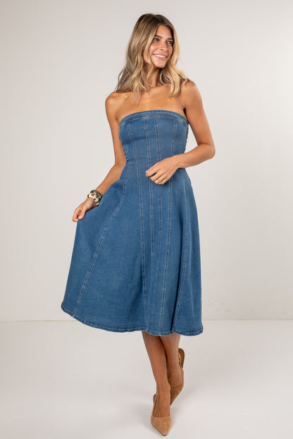 A woman modeling a strapless blue denim midi dress with a flared skirt and front pockets, paired with tan pointed-toe heels against a plain light gray studio background.