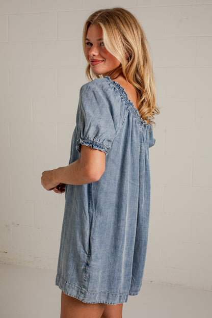 A woman wearing a light blue denim short-sleeve dress with ruffled trim, photographed against a white brick wall.