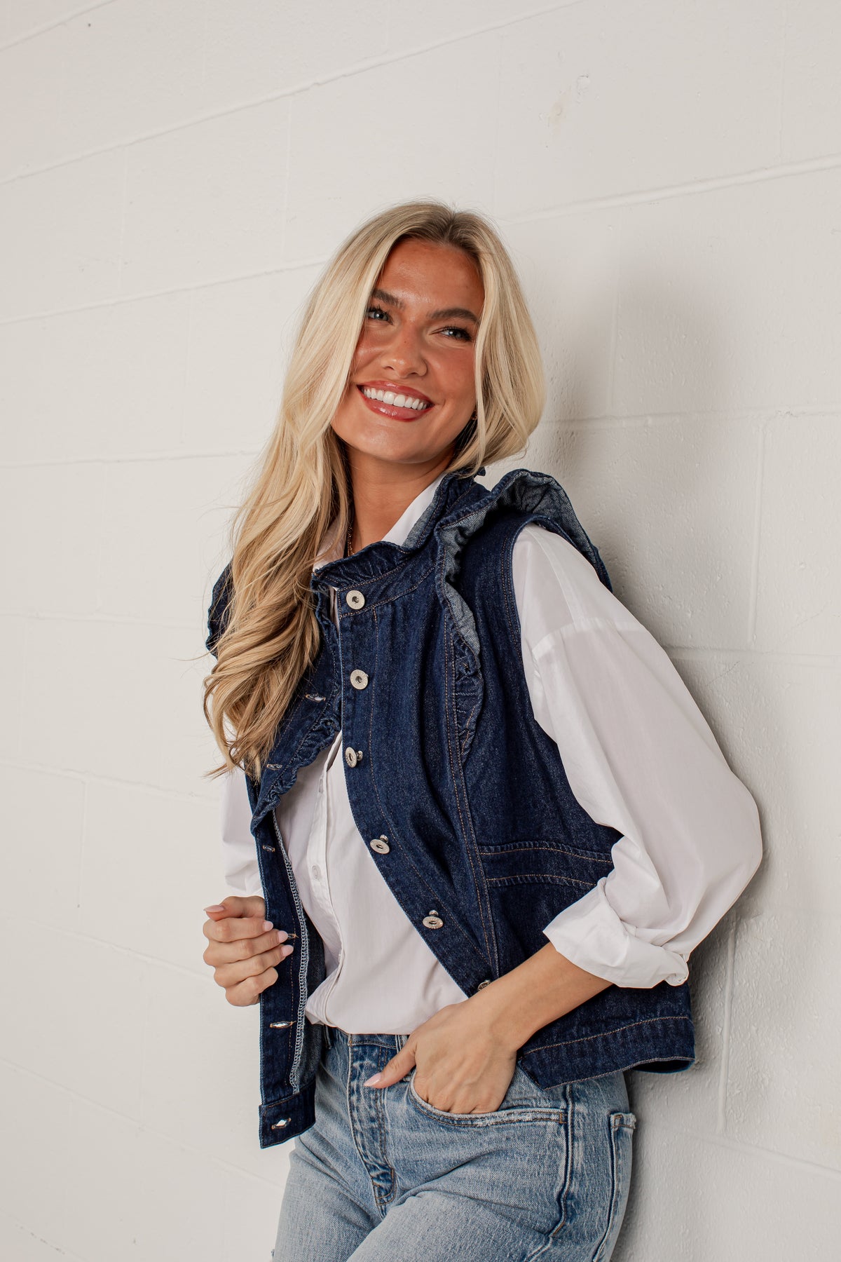A smiling blonde woman leans against a white paneled wall wearing a dark blue denim vest over a white long-sleeve blouse and light blue jeans.