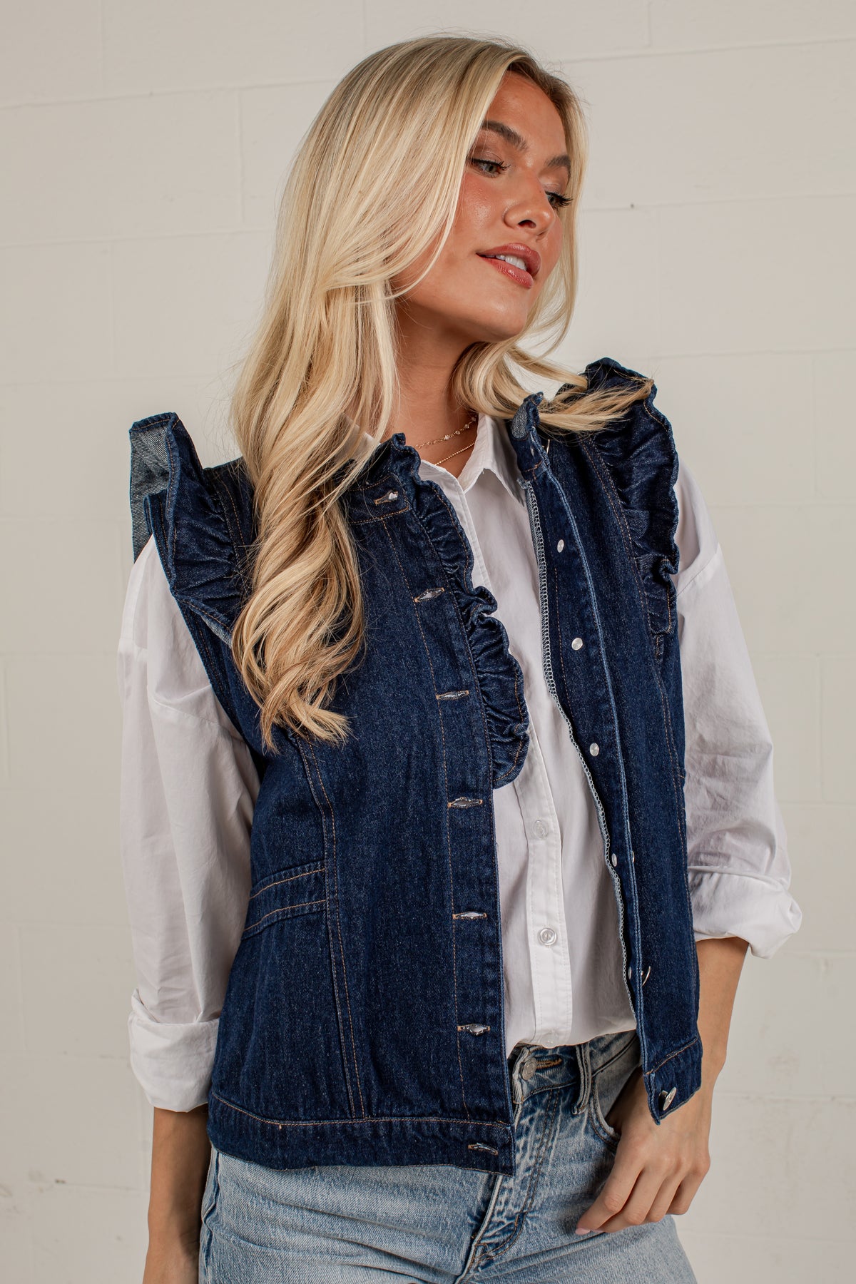 A blonde woman wearing a dark blue denim vest with ruffle trim over a white button-up shirt and light blue jeans, standing against a light-colored brick wall.
