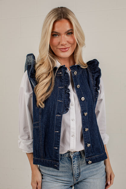 A blonde woman wearing a dark blue denim vest with ruffle trim over a white button-up shirt and light blue jeans, standing against a light off-white brick wall.