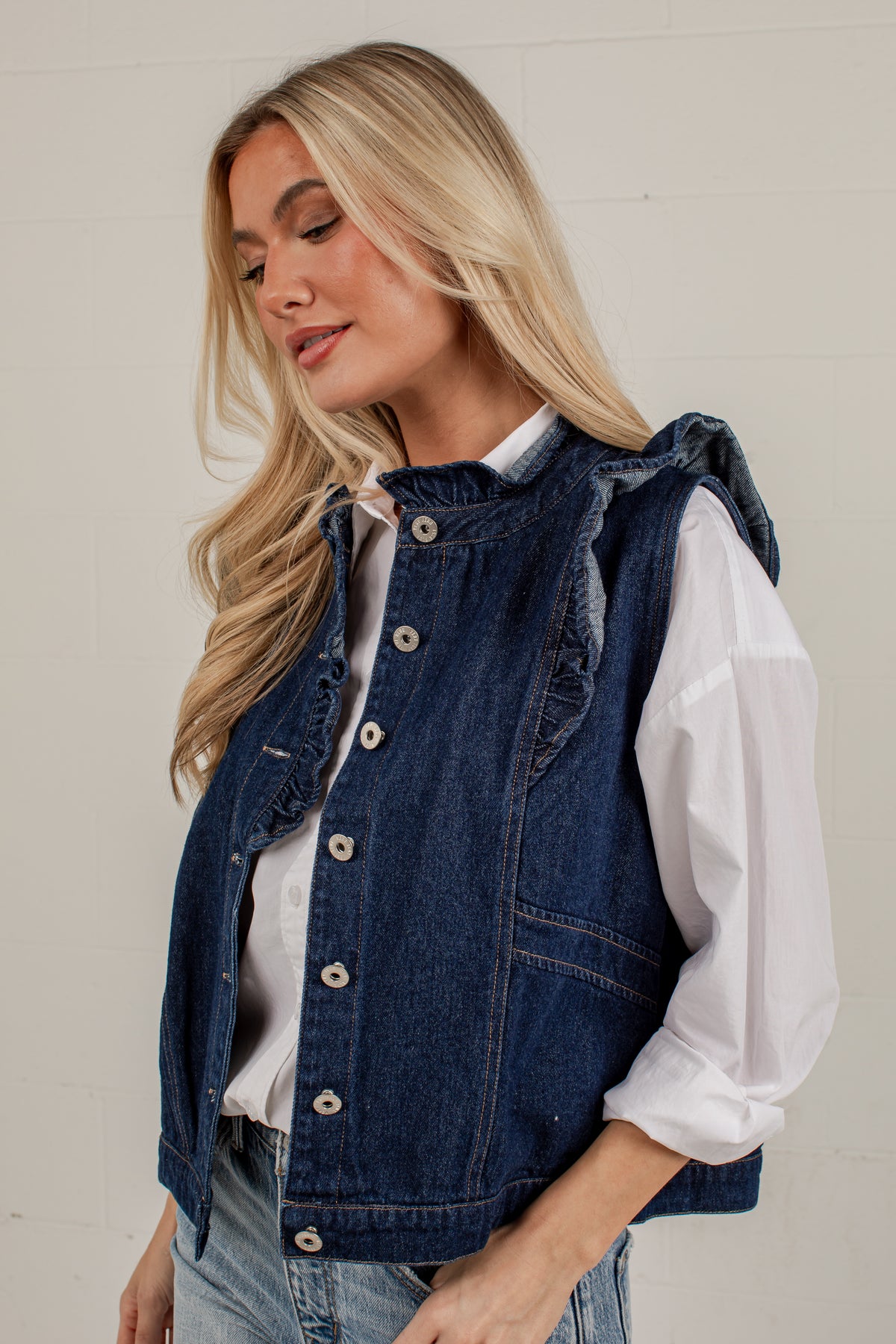 A blonde woman wearing a dark blue denim sleeveless vest over a white long-sleeve shirt and light blue jeans, standing against a light off-white paneled wall.