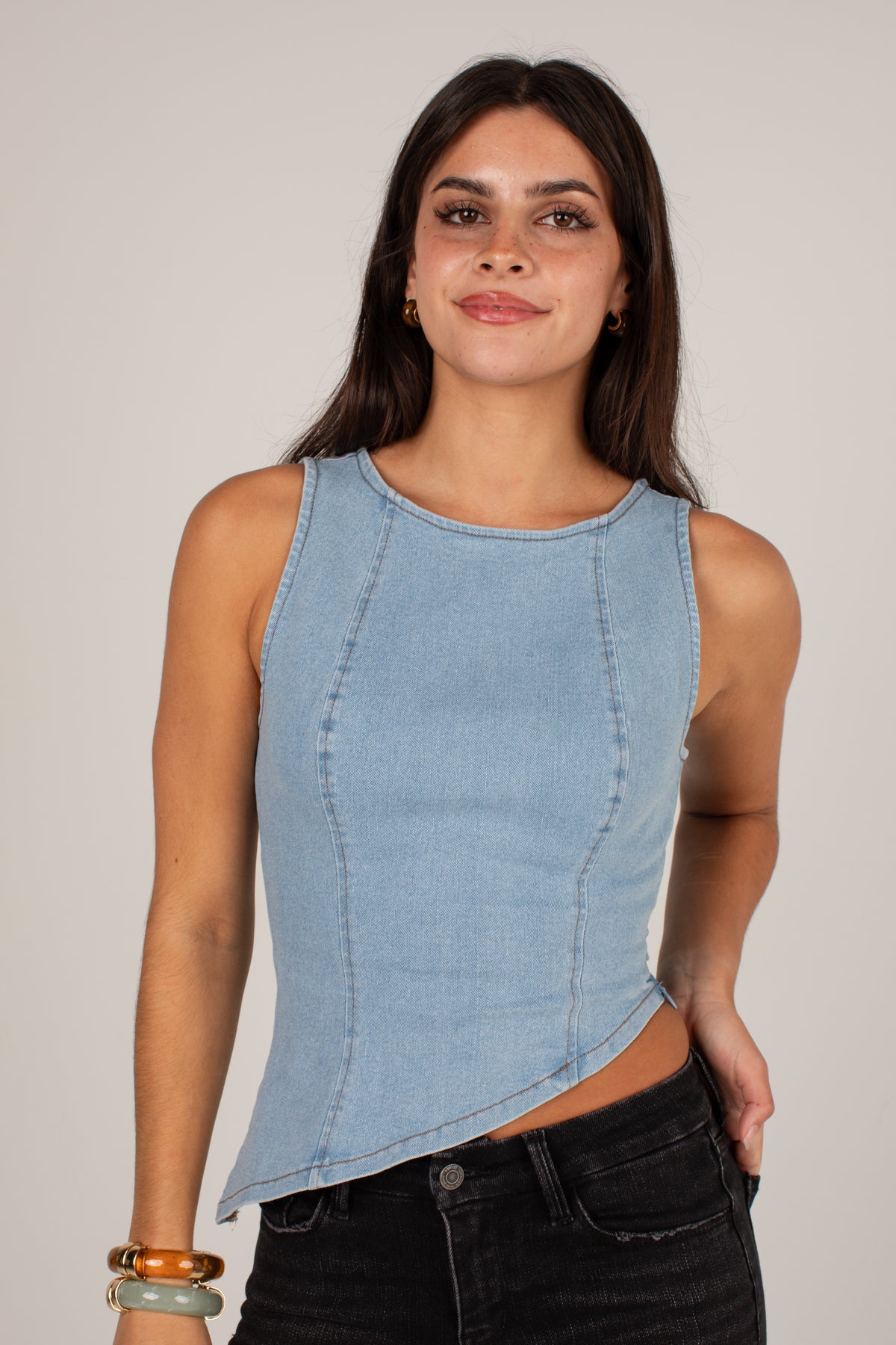 A woman modeling a sleeveless light blue denim top with a curved asymmetrical hem, paired with black jeans, against a plain light gray studio background.