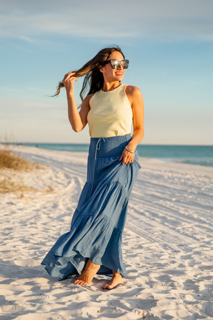 Woman in a yellow top and blue skirt standing on a sandy beach with ocean and sky in the background.
