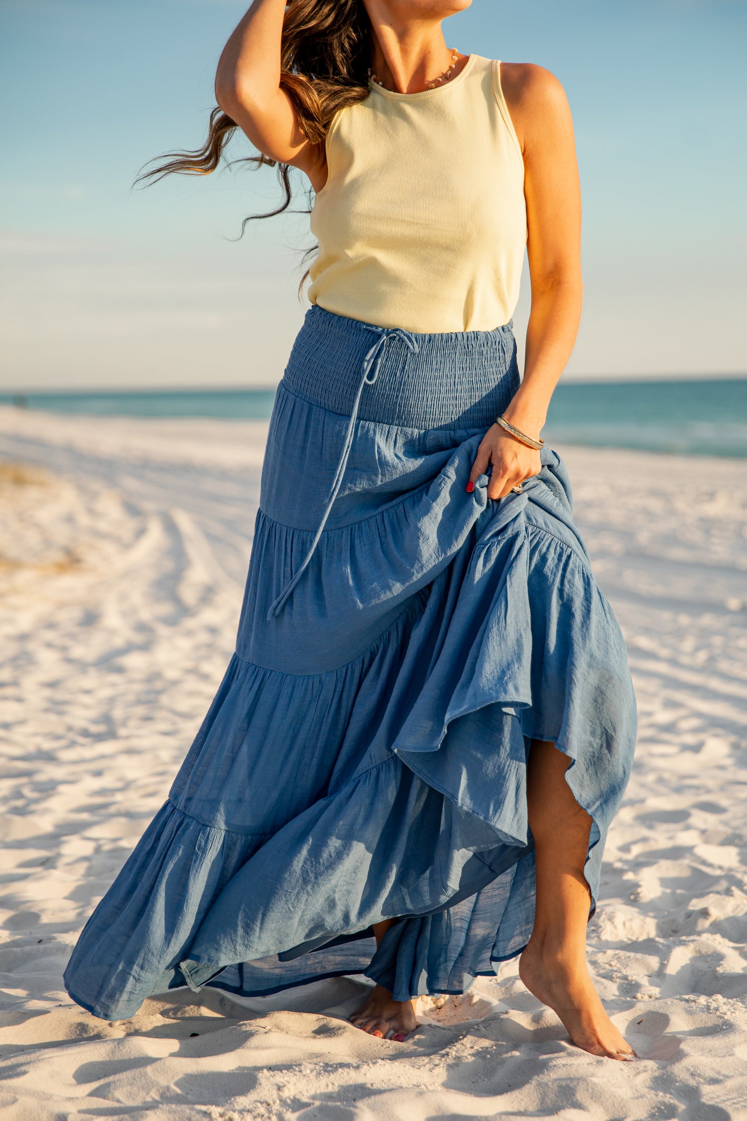 Woman in a yellow top and blue denim skirt standing on a sandy beach.
